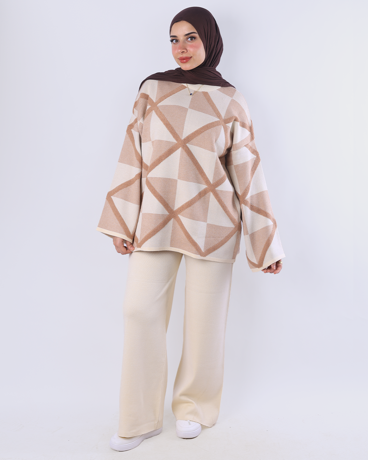 Woman wearing a beige and brown geometric patterned top with white pants on a white background