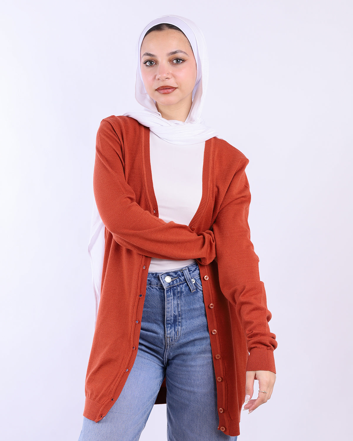 Woman wearing a rust-colored cardigan, white top, and blue jeans on a white background- WIND Shopping